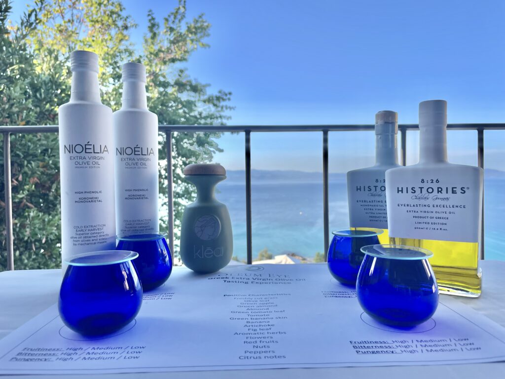 Bottles of olive oil with blue tasting glasses and a tasting chart on a table overlooking a scenic sea view.
