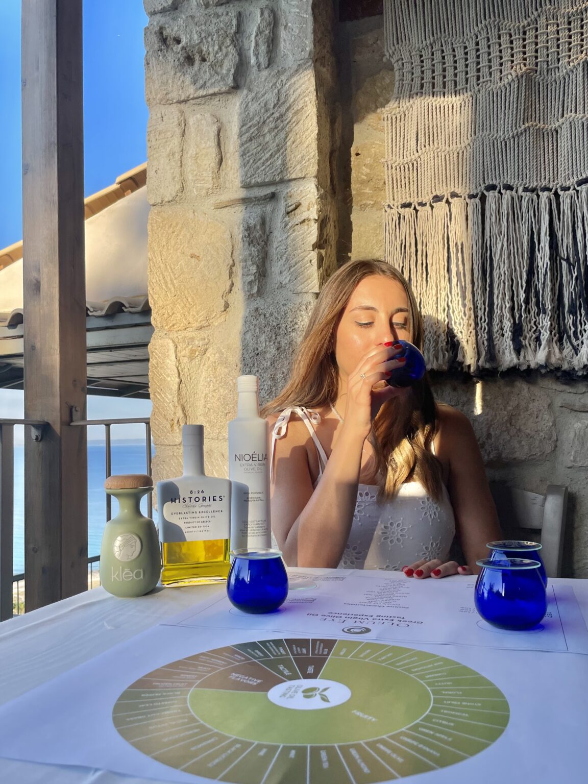 Woman tasting olive oil at an olive oil tasting event with bottles of olive oil and a tasting chart on the table.