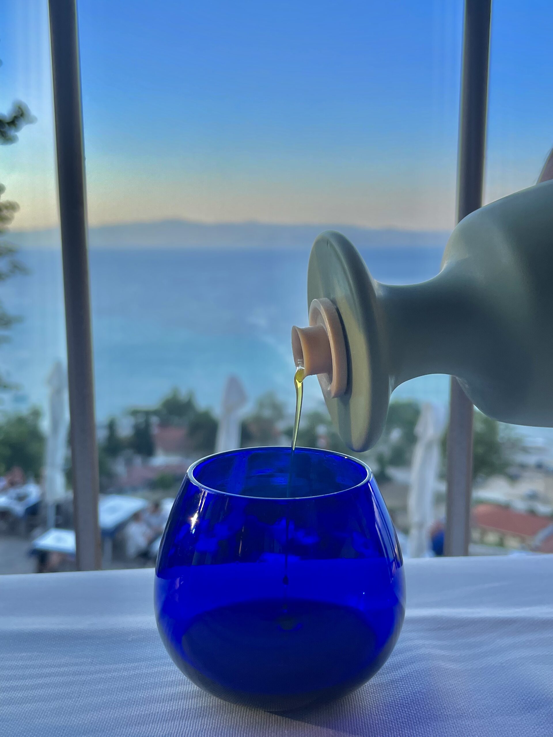 Olive oil being poured into a blue tasting glass with a scenic sea view in the background.